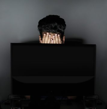 man appearing stressed looking at a laptop screen with hands over eyes.